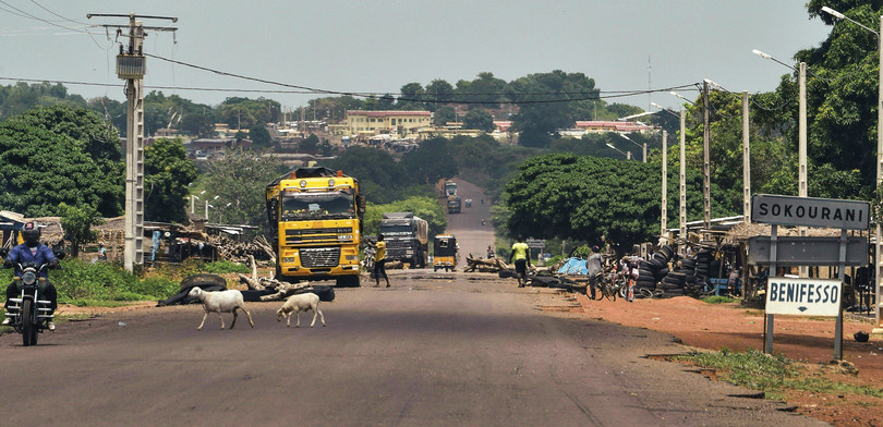 View down a tarred road with dirt pavements, crossed by aerial power lines. A man rides a motocycle, two sheep cross the road, pedestrians walk beside the road. A large yellow truck drives towards us.
