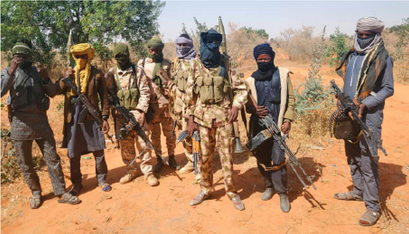 A group of eight men with rifles, some in military camouflage, with their faces covered, looking towards us posing for this photograph. They are outdoors on sandy ground, with scrub and trees in the background.
